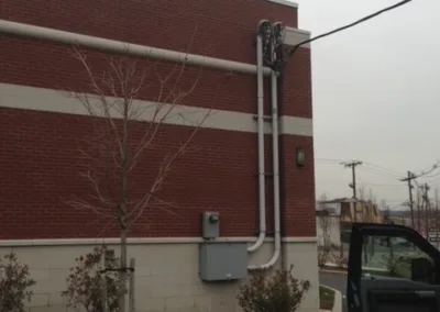 An external view of a building with electrical conduit and utility boxes on the brick wall, with shrubs and a leafless tree in the foreground.