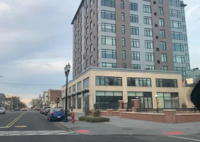 A modern multistory building on a street corner with cars parked along the roadside, viewed from inside a car.