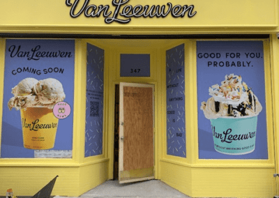 Storefront of Van Leeuwen, a new ice cream shop opening soon, with yellow façade, large window posters of ice cream cones, and a text "Good for you. Probably." at 347 address. Orange cone on the sidewalk.