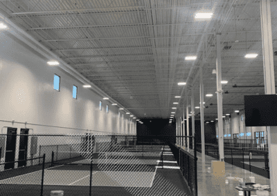 A large, empty indoor facility with high ceilings, white walls, and bright overhead lights. The floor is concrete, and a black chain-link fence encloses several areas. Containers and tools sit on the floor.