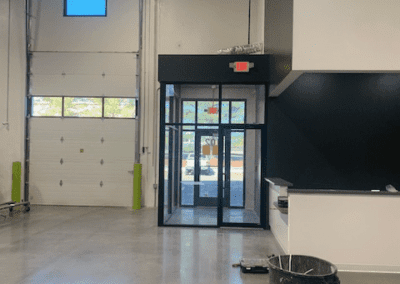 A spacious indoor area with polished concrete floors, a large roll-up door, a revolving door, and a black accent wall. A trash bin, boxes, and cleaning supplies are scattered on the floor.