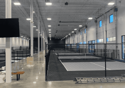 A spacious indoor facility with multiple sections divided by fencing, polished concrete floors, high ceilings, and ample lighting. Benches and a large TV screen are visible in the foreground.