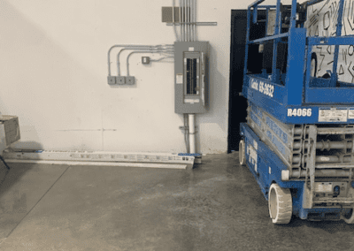 A blue scissor lift is parked indoors near a white wall. The wall has multiple electrical conduits and a breaker panel. The concrete floor appears newly cleaned.