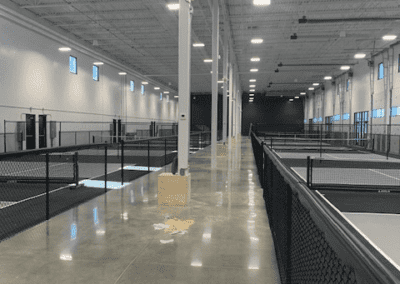 A large, empty indoor warehouse with a polished concrete floor, high ceiling, overhead lights, and chain-link fences dividing sections of the space.