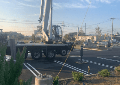 A crane truck is parked on a paved surface in a parking lot, with its boom extended upwards. A nearby concrete block and a bush are in the foreground. Buildings and power lines are in the background.