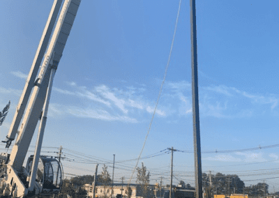 A man is guiding the installation of a tall streetlight using a crane in a parking lot. The weather is clear with blue skies.