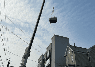 A crane lifts a large object near multi-story buildings under a partly cloudy sky. Power lines and residential houses are visible in the foreground.