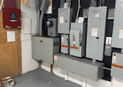 A room with electrical panels mounted on the wall, various wires connected to them, and a red fire alarm control panel to the left. The floor is gray and clean, and the ceiling has exposed beams and pipes.