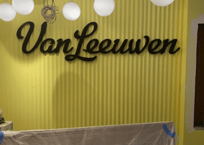 A yellow wall with the black "Van Leeuwen" sign. Ceiling lights are hanging, and a workbench with tools and a covered bench are in the foreground.