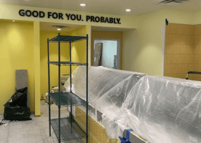 A room under construction with yellow walls, plastic sheets covering surfaces, and a metal shelving unit in the center. The wall has a sign that reads "GOOD FOR YOU. PROBABLY.