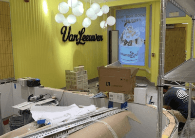 Interior view of a Van Leeuwen store under construction with boxes, construction materials, and tools scattered around the partially finished space and a neon Van Leeuwen sign on the bright yellow wall.