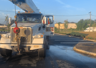 A truck with an extended crane arm is parked on a paved surface under a clear sky. A person is visible near the open driver-side door. There is some water or liquid visible on the pavement.